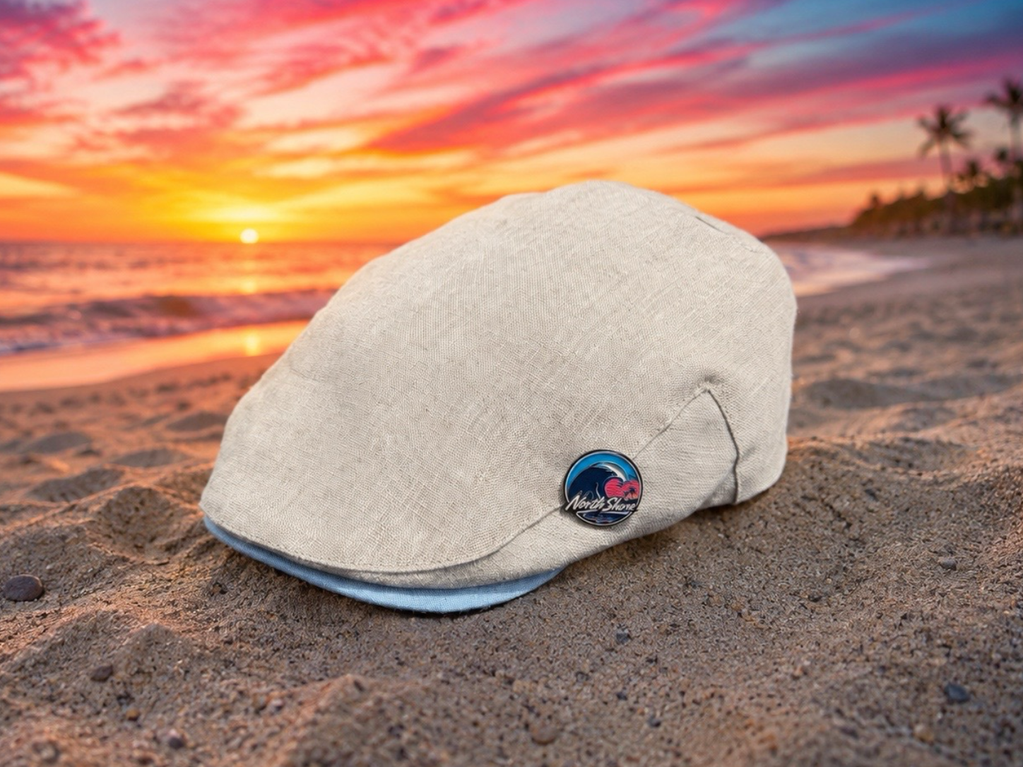 A light beige flat cap resting on a sandy beach with a sunset ocean backdrop. The cap features a small circular blue and red enamel pin on the side.