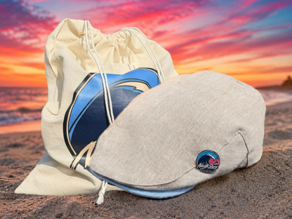 A light beige linen cap with a circular blue and red wave logo enamel pin resting on a sandy beach in front of a matching beige drawstring bag with a large blue wave graphic, set against a vibrant sunset sky.