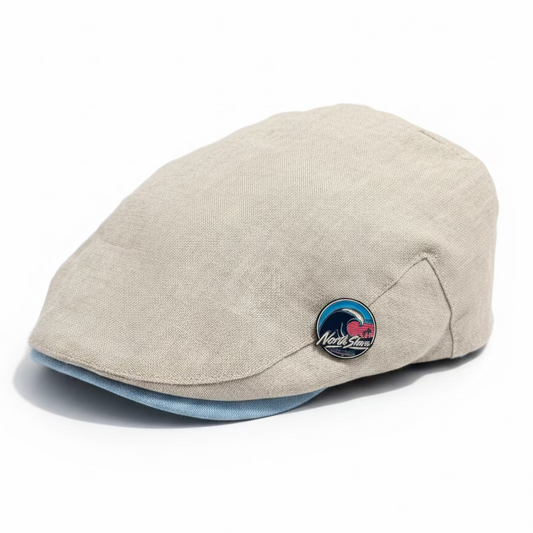 A light beige linen flat cap with a light blue visor underside and a circular enamel pin on a white studio background 
