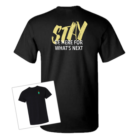 A black short-sleeve T-shirt shown from the back with a large graphic print reading “STAY BE HERE FOR WHAT’S NEXT” in gold and white. A smaller inset view in the lower left shows the front of the same shirt with a tiny green and red chest graphic.
