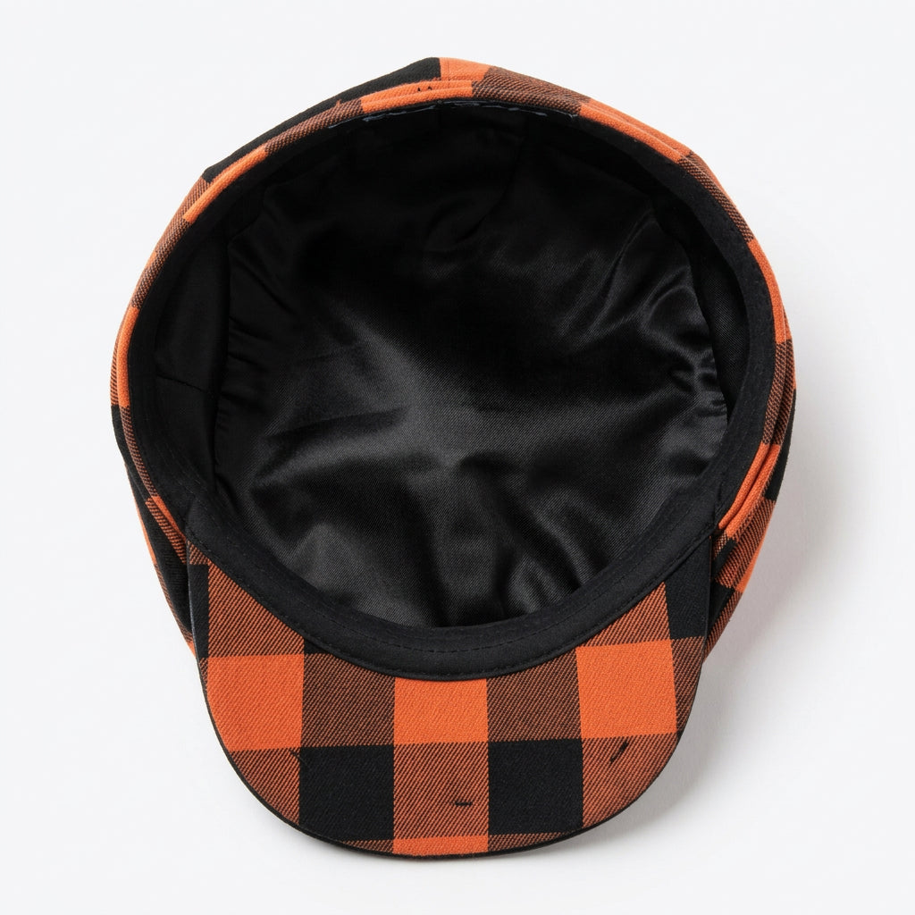 A top-down view of a black and orange buffalo plaid flat cap resting on a light plain white background 