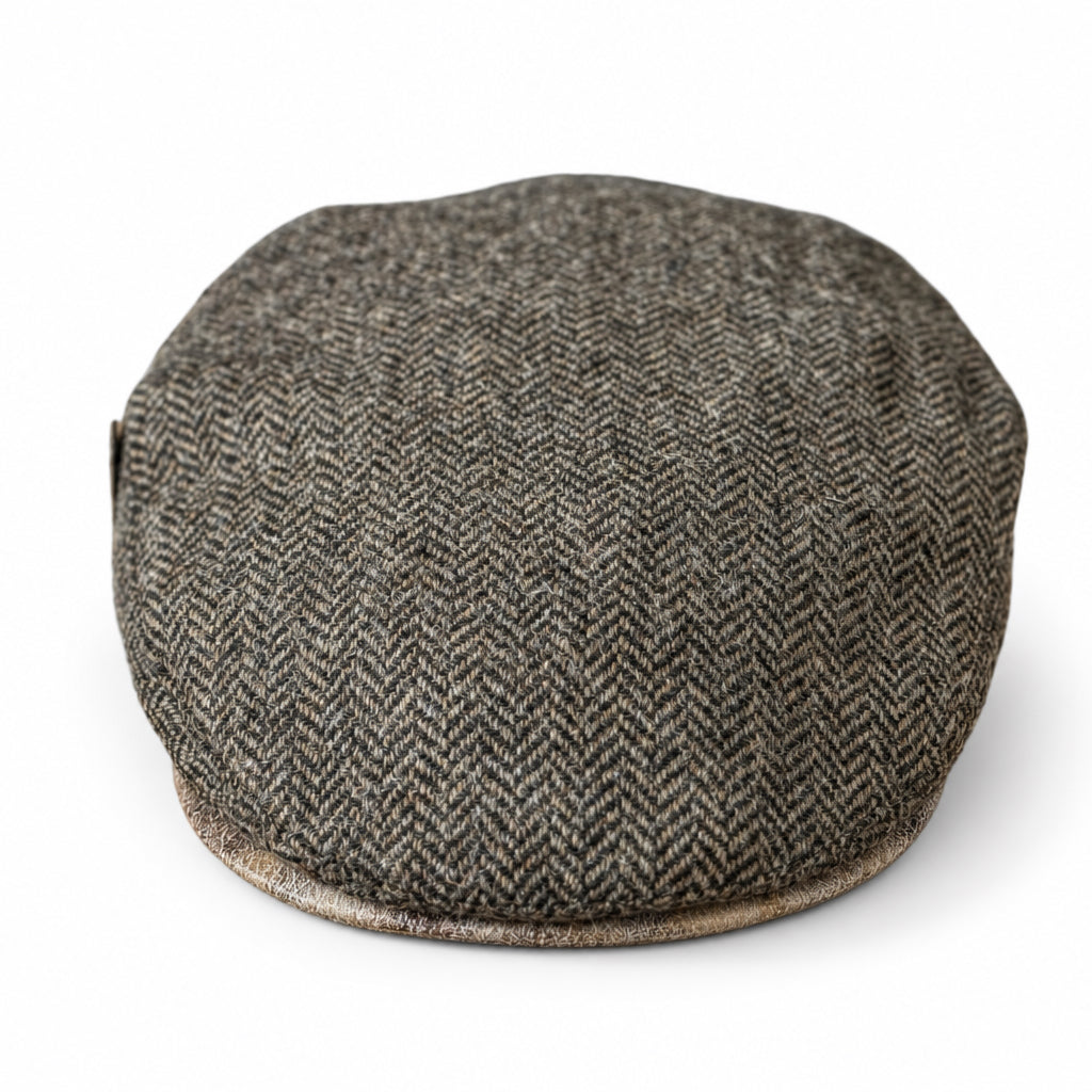 A brown herringbone tweed flat cap resting with a light contrasting brim on a solid white background 